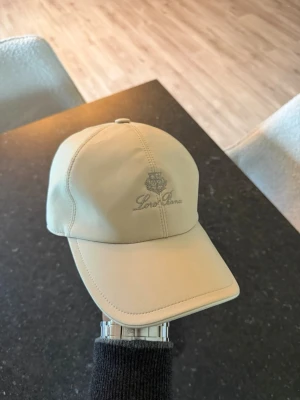 Loro Piana keps  - Snygg beige keps från Loro Piana med broderad logga framtill. Klassisk böjd skärm och sex paneler med synliga sömmar. Tillverkad i ett slätt tyg som ger en stilren look. Perfekt accessoar för en clean och avslappnad stil.
