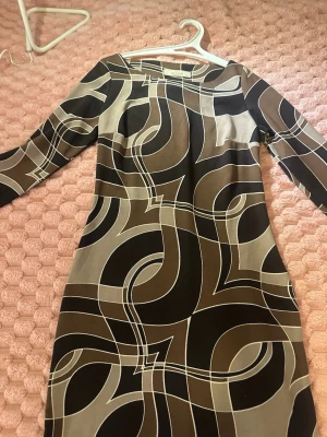 Mönstrad klänning från Salve - Cool klänning från Salve med grafiskt mönster i brunt, beige och svart. Klänningen har långa ärmar och en rak passform. Materialet känns mjukt och stretchigt, perfekt för dig som gillar retrovibbar och unika prints.