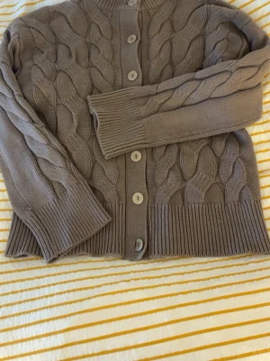 Brun kabelstickad kofta 157 XS - Mysig brun kabelstickad kofta från 157 med stora knappar framtill. Koftan har ribbade muddar och nederkant samt ett klassiskt flätmönster över hela plagget. Perfekt för lager på lager och en chill look.passar perfekt nu till hösten!🥰