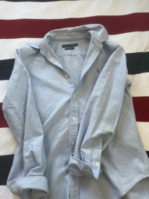 Ljusblå skjorta från Ralph Lauren - Klassisk ljusblå skjorta från Ralph Lauren med knappstängning och broderad logga på bröstet. Skjortan har lång ärm, krage och är tillverkad i mjuk bomull. Perfekt för en clean och stilren look.