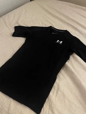 Svart kompressionströja Under Armour - Säljer en svart kompressionströja från Under Armour med HeatGear-teknologi. Tröjan har korta ärmar, rund hals och en vit logga på bröstet. Perfekt för träning och sportaktiviteter där du vill hålla dig sval och bekväm.