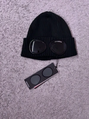 Svart mössa med goggles från C.P. Company - Säljer en svart ribbstickad mössa från C.P. Company med ikoniska goggles på uppviket. Mössan är tillverkad i mjukt material och har en modern streetwear-look. Perfekt accessoar för att lyfta din outfit.