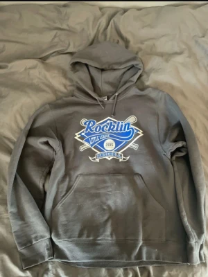 Grå vintage hoodie med Rocklin Little League-tryck - Säljer en grå hoodie med stor Rocklin Little League Baseball-logga i blått och vitt på bröstet. Hoodien har huva med dragsko och en klassisk magficka. Perfekt för dig som gillar sportig stil och vill ha något bekvämt och snyggt. Köpt i Stocholm för 300.