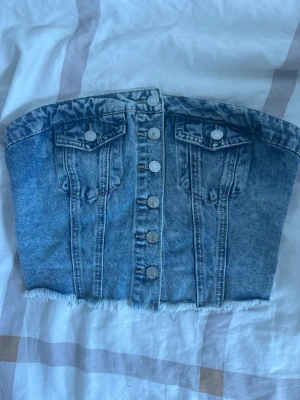 Blå jeans tubtopp med knappar - Säljer en snygg blå tubtopp i jeanstyg med knappar framtill och två dekorativa bröstfickor. Toppen har rå kant nertill för en trendig look och är perfekt för dig som vill sticka ut.