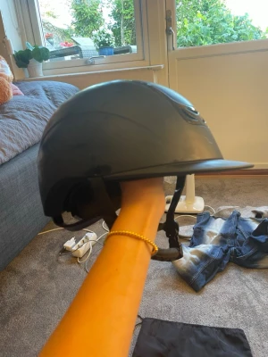 Svart ridhjälm från JH Collection - Snygg svart ridhjälm från JH Collection med blank finish och ventiler upptill. Hjälmen har justerbart hakband och kommer med en praktisk förvaringspåse. Perfekt för dig som vill ha både stil och säkerhet i stallet.