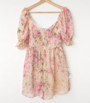 Blommig kortklänning från Miss Selfridge - Ny oanvänd! Romantisk kortklänning (med shorts under) från Miss Selfridge i ljusa pastellfärger med blommigt mönster. Klänningen har puffärmar, spetsdetaljer vid urringningen och knappar framtill. Perfekt för sommarens alla tillfällen.