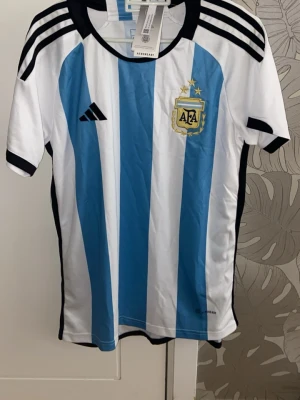 Argentina fotbollströja - Säljer en officiell Argentina fotbollströja från Adidas med klassiska blå och vita vertikala ränder. Tröjan har svarta detaljer, AFA-emblem på bröstet och tre svarta ränder på axlarna. Tillverkad i lätt och ventilerande material, perfekt för träning eller match.