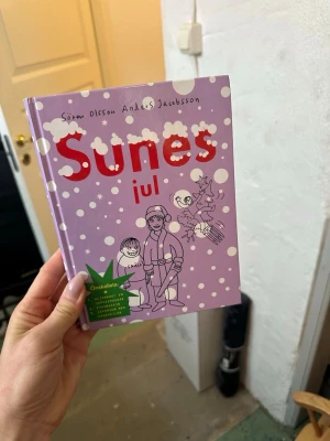 Sunes jul av Sören Olsson - Rolig och vintrig bok med lila hård pärm, snöbollsmotiv och illustrationer på framsidan. Boken är skriven av Sören Olsson och Anders Jacobsson och handlar om Sunes juläventyr. Perfekt för dig som gillar humor och julstämning.