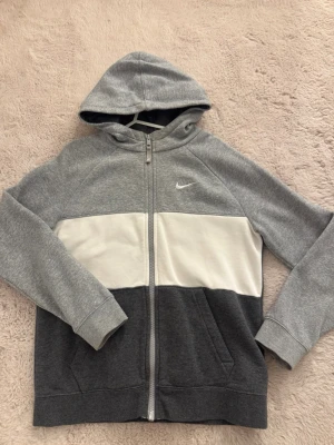 Nike hoodie - Snygg hoodie från Nike med dragkedja, huva och fickor framtill. Tröjan har färgblock i ljusgrått, vitt och mörkgrått samt Nike-logga broderad på bröstet. Tillverkad i mjukt sweatshirtmaterial som är perfekt för chill dagar. (Lappen är tyvärr bortklippt så vet ej vilken storlek, men skulle gissa på M/L) 💗💗💗
