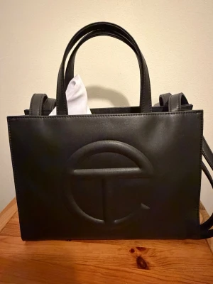 Svart handväska från Telfar - Stilren svart handväska från Telfar i rektangulär form med präglad logga på framsidan. Väskan har dubbla handtag och en lång axelrem för olika bärstilar. Tillverkad i veganskt läder och kommer med en vit dustbag med stor logga. 