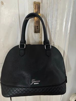 Svart handväska från Guess - Snygg svart handväska från Guess med quiltad botten och två rejäla handtag. Väskan har en rundad form och är tillverkad i konstläder med silverfärgade detaljer. Guess-logga framtill ger en exklusiv touch. Perfekt för dig som gillar stilrena accessoarer.