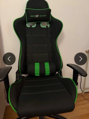 Svart och grön gamingstol - Säljer en gamingstol i svart med gröna detaljer och vita sömmar. Stolen har justerbara armstöd, nackkudde och svankstöd. Märket 'apeX racer' syns på nackkudden. Perfekt för långa spelsessioner eller hemmakontoret.