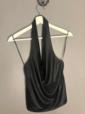 Svart halterneck topp i satin - Snygg svart halterneck topp i glansigt satinmaterial med draperad front och tunna axelband. Perfekt för dig som vill ha en trendig och elegant look. Toppen är ärmlös och har en croppad passform.
