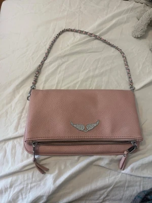 Rosa kuvertväska med kedjerem - Snygg rosa kuvertväska i skinnimitation med silverfärgad kedjerem och dekorativt emblem med vingar framtill. Väskan har dragkedja både på framsidan och upptill samt beige innerfoder. Perfekt storlek för det viktigaste och enkel att bära över axeln.