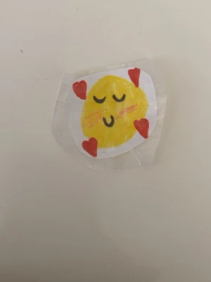Handgjord emoji-klistermärke med hjärtan - Unikt handgjort klistermärke med en gul emoji som har stängda ögon och fyra röda hjärtan runt sig. Motivet är ritat på vitt papper och täckt med tejp för extra hållbarhet. Perfekt att dekorera dator, mobil eller anteckningsbok med.