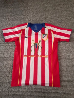 Atletico Madrid 04/05 Spider-Man matchtröja - Röd och vitrandig Atletico Madrid matchtröja från Nike med blå detaljer. Framsidan har ett stort Spider-Man 2-tryck och klubbmärke på bröstet. Tillverkad i lätt och ventilerande material, perfekt för fotboll eller träning.