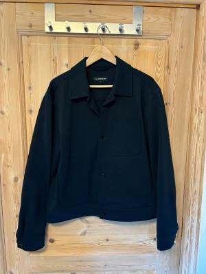 Mörkblå overshirt från J.Lindeberg - Stilren mörkblå overshirt från J.Lindeberg med klassisk krage, knappstängning och en bröstficka. Overshirten har raka ärmar med knapp vid ärmslut och är tillverkad i ett kraftigare tyg som ger en snygg och modern look.