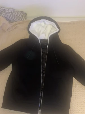Moose knuckles kofta  - Säljer en svart hoodie med dragkedja och vit fleecefoder i huvan. Hoodien har en stor huva med dragsko och en unik metall-logga på ärmen. Perfekt för dig som vill ha något både snyggt och bekvämt. Materialet känns mjukt och varmt.