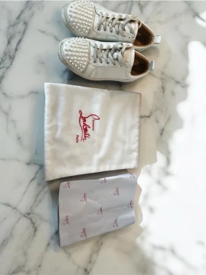 Vita sneakers med nitar från Louboutin - Säljer ett par vita sneakers från Christian Louboutin med silverfärgade nitar på tån och den ikoniska röda sulan. Skorna har vita skosnören och detaljer i läder. Medföljer originalpåse och papper.