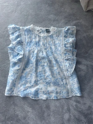 Blommig volangblus från Gina Tricot - Supersöt blus från Gina Tricot i vitt med blått blommönster. Blusen har volangdetaljer längs ärmarna, spets vid halsen och fina veck framtill. Perfekt för dig som gillar romantisk och feminin stil. (Har bara använt en gång)
