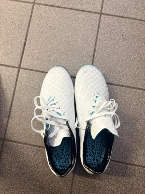 Vita fotbollsskor med blå detaljer - Säljer ett par vita Nike fotbollsskor med blå dobbar och mönstrad insida. Skorna har snörning, svart detalj vid hälen och en modern, sportig look. Ovandelen är i syntetmaterial med struktur för bättre bollkontroll. Perfekta för dig som vill sticka ut på planen! Vid snabb affär sänks priset!
