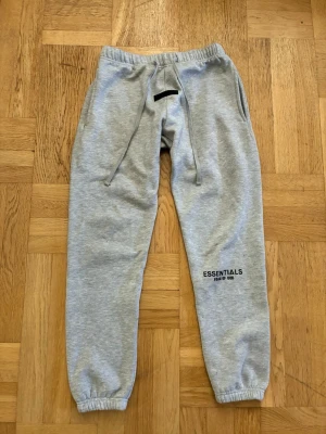 Essentials mjukisbyxor - Snygga grå mjukisbyxor från Fear of God Essentials med resår i midjan och benslut, dragsko och fickor på sidorna samt en bakficka. Logga tryckt på vänster ben. Perfekta för chill dagar och streetwear-stil.