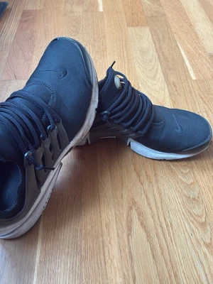 Svarta Nike sneakers med vit sula - Säljer ett par svarta Nike sneakers med vit sula och svart mesh-överdel. Skorna har en plastdetalj på sidan och klassisk Nike-logga. Lågt skaft och snörning framtill. Perfekta för en sportig och avslappnad stil. Denna model säljs inte längre vilket gör den sällsynt. Ena skon har dock ett litet hål vilket gör att jag säljer för väldigt billigt. 