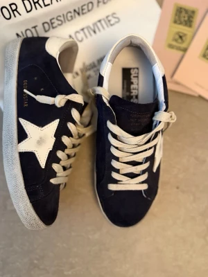Golden Goose svarta sneakers med stjärna - Snygga mörkblå sneakers från Golden Goose med vit stjärndetalj på sidan och vit hälkappa. Skorna har vita skosnören och en slitstark sula. Ovandelen är i mocka och detaljerna i läder. 
