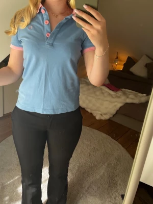 Blå pikétopp med rosa detaljer Fred Perry - Snygg blå pikétopp från Fred Perry med rosa krage, knappar och ärmslut. Klassisk broderad logga på bröstet. Helt oandvänd och nypris 1000kr-1400kr