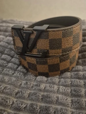 Brunt rutigt bälte med svart spänne - Snyggt bälte med klassiskt rutigt mönster i brunt och svart samt ett stort spänne i svart metall. Perfekt accessoar för att lyfta din outfit och ge en exklusiv touch. Bältet har flera hål för justerbar passform.