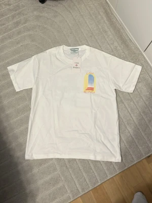 Vit Casa Blanca t-shirt med tryck - Vit t-shirt från Casa Blanca med färgglatt tryck framtill som föreställer en öppen dörr mot himlen. Klassisk rund hals och korta ärmar. Tillverkad i mjuk bomull, perfekt för en avslappnad och stilren look.