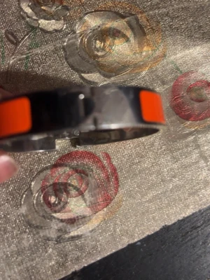 Orange och silverfärgat armband - Snyggt armband i metall med en bred orange emaljdetalj runt hela bandet. Armbandet har en stilren och modern design med blanka silverfärgade partier och ett smidigt spänne. Perfekt för dig som vill ha en färgklick till din outfit.