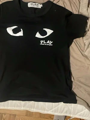 Svart Comme des Garçons PLAY t-shirt - Säljer en svart t-shirt från Comme des Garçons PLAY med vita ögon och logga tryckt på bröstet. Klassisk rund hals och korta ärmar. 