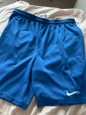 träningsshorts - Snygga blå shorts från Nike med vit logga på benet och praktisk snörning i midjan. Perfekta för träning eller sportiga dagar. De har en klassisk passform och är gjorda i ett lätt och bekvämt material.