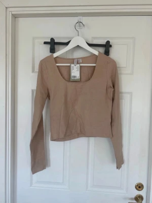 Beige långärmad croppad topp - Trendig beige croppad topp med lång ärm och djup rundringning. Tillverkad i mjukt bomullsmaterial som känns skönt mot huden. Perfekt att styla med höga jeans eller kjol för en clean look.
