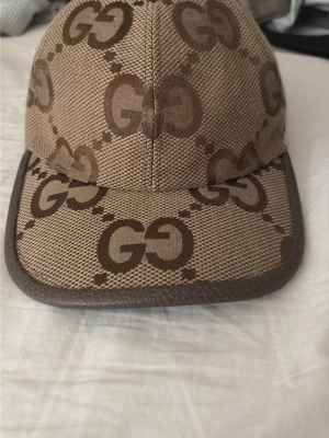 Beige keps med GG-mönster - Snygg beige keps med brunt GG-mönster över hela kepsen och brun kant runt skärmen. Tillverkad i ett tåligt tygmaterial och har en klassisk form med böjd skärm. Perfekt för dig som gillar stilrena accessoarer med logga.