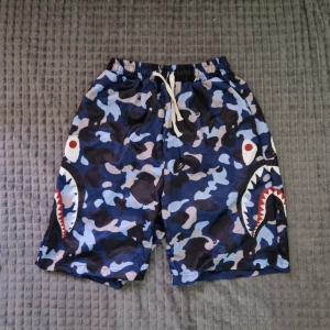Blå BAPE med hajmotiv - Säljer ett par blå camouflagemönstrade shorts med hajmotiv på sidorna. Shortsen har resårmidja med vit snörning och är tillverkade i ett lätt syntetmaterial. Perfekta för dig som vill sticka ut med en unik och streetinspirerad stil.