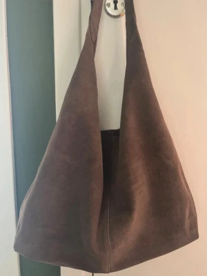 Brun shoppingväska i mocka - Stilren shoppingväska i mjuk brun mocka med enkel, rymlig design och breda axelband. Väskan har en avslappnad form och passar perfekt för dig som gillar minimalistisk stil. Perfekt för att bära allt du behöver under dagen.