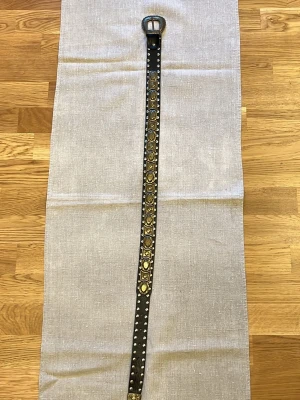 Svart bälte med nitar och dekorationer 94cm - Snyggt svart bälte med silverfärgat spänne och massor av runda nitar samt guldfärgade dekorationer längs hela bältet. Perfekt för att ge outfiten en edgy touch. Bältet är smalt och har en cool, rockig vibe.