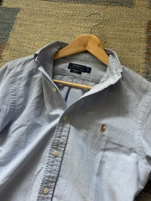 Ljusblå skjorta från Polo Ralph Lauren - Klassisk ljusblå skjorta från Polo Ralph Lauren med broderad logga på bröstet. Skjortan har lång ärm, knappar framtill och en tidlös design i bomull. Perfekt för en clean och stilren look.