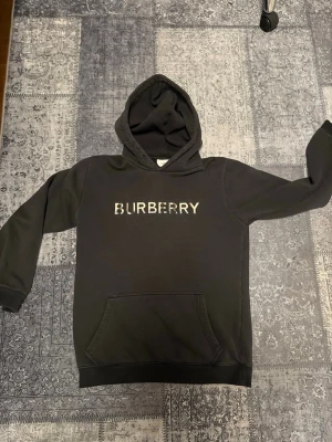 Svart B hoodie med logga - Säljer en svart hoodie från B med stor broderad b på Hoodien, den har huva och en klassisk magficka. Perfekt för dig som gillar streetwear och vill ha något stilrent men ändå lyxigt.