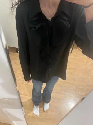 Svart blus med volangdetaljer - Snygg svart blus med stora volangdetaljer framtill och klassisk krage. Blusen har knäppning hela vägen och långa ärmar. Passar perfekt till jeans eller kjol för en trendig look. Materialet ser ut att vara mjukt och följsamt.
