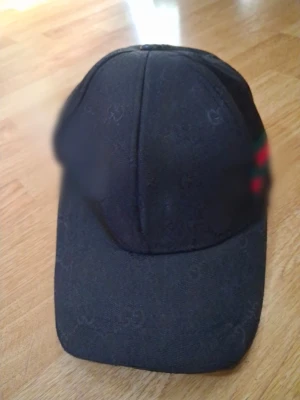 Svart keps med diskret GG-mönster - Snygg svart keps med diskret GG-mönster över hela ytan och klassisk böjd skärm. På sidan finns en detalj med gröna och röda ränder. Kepsen har en stilren och modern look, perfekt för att lyfta din outfit.