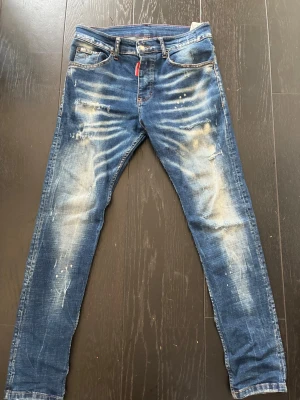 Blå slitna jeans från Dsquared2 - Säljer ett par blå jeans från Dsquared2 med slitningar, blekta partier och färgstänk för en riktigt trendig look. Jeansen har smal passform, klassisk femficksmodell och knappgylf. Perfekta för dig som gillar streetwear och en edgy stil. PRIS går att diskuteras!