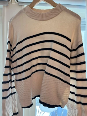 Randig stickad tröja från Gina Tricot - Säljer en vit stickad tröja från Gina Tricot med svarta horisontella ränder. Tröjan har ribbad rund halsringning, långa ärmar och ribbade muddar. Perfekt för dig som gillar klassisk och stilren look.