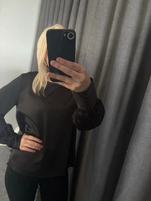 Svart satinblus från Vero Moda - Stilren svart blus i satin från Vero Moda. Blusen har lång ärm med knappar vid ärmslut och en rak passform. Materialet är glansigt och ger en lyxig känsla. Perfekt att styla med jeans eller kjol för en trendig look.