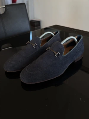 By Hudson mocka loafers - Snygga mörkblå loafers i mocka med elegant metallspänne över vristen. Skorna har en klassisk, smal siluett och låg klack. Perfekta för dig som gillar stilrena detaljer och en dressad look. Yttermaterialet är mjukt och ger en exklusiv känsla.