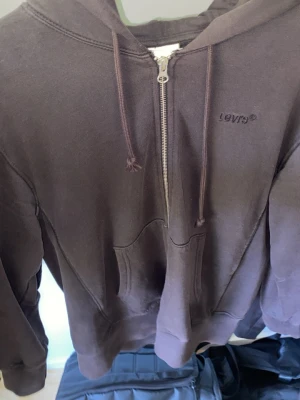 Brun zip hoodie från Levi's - Säljer en brun hoodie från Levi's med dragkedja framtill, huva med dragsko och känguruficka. Klassisk och enkel design med Levi's-logga broderad på bröstet. Perfekt för chill dagar och streetwear-stil.