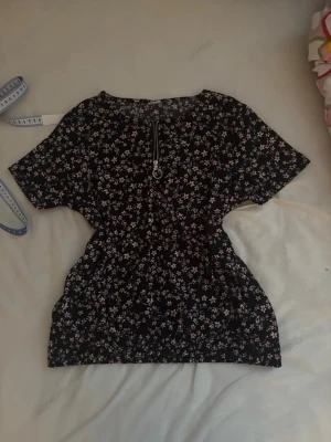 Svart blommig topp - Söt svart topp med små blommor i vitt och rosa. Toppen har korta ärmar och en dekorativ dragkedja framtill med ringdetalj. Perfekt för dig som gillar en feminin och avslappnad stil.