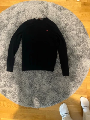 Mörkblå stickad tröja från Morris - Snygg mörkblå stickad tröja från Morris med rund halsringning och diskret röd logga broderad på bröstet. Tröjan har långa ärmar och ribbade muddar vid ärmslut och nederkant. Perfekt till lager på lager och enkel att matcha. Är i storlek S men sitter mer som en xs då de är små i storlekarna.